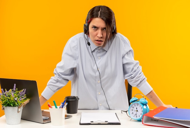 Garota jovem descontente do call center usando fone de ouvido, levantando-se da mesa com ferramentas de trabalho isoladas em um fundo laranja