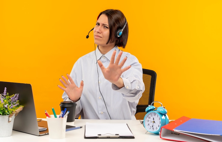 Foto grátis garota jovem descontente do call center usando fone de ouvido, sentada na mesa com as ferramentas de trabalho, sem fazer nenhum gesto isolado em um fundo laranja