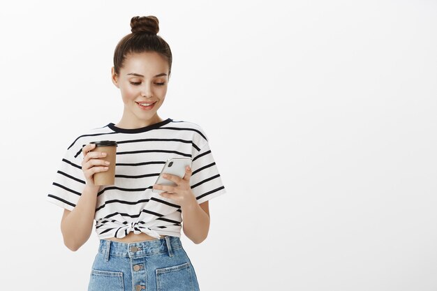 Garota jovem e atraente usando smartphone enquanto bebe café, segurando a xícara e o telefone celular