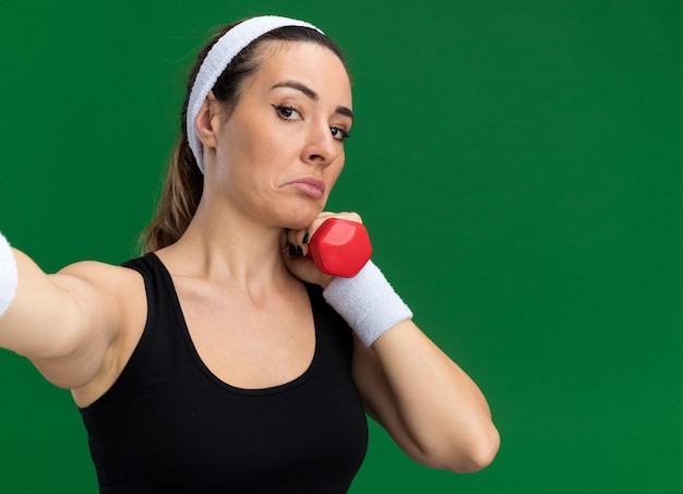 Garota jovem e bonita e confusa, esportiva, usando bandana e pulseiras, segurando um haltere, estendendo a mão em direção à câmera, isolada na parede verde com espaço de cópia