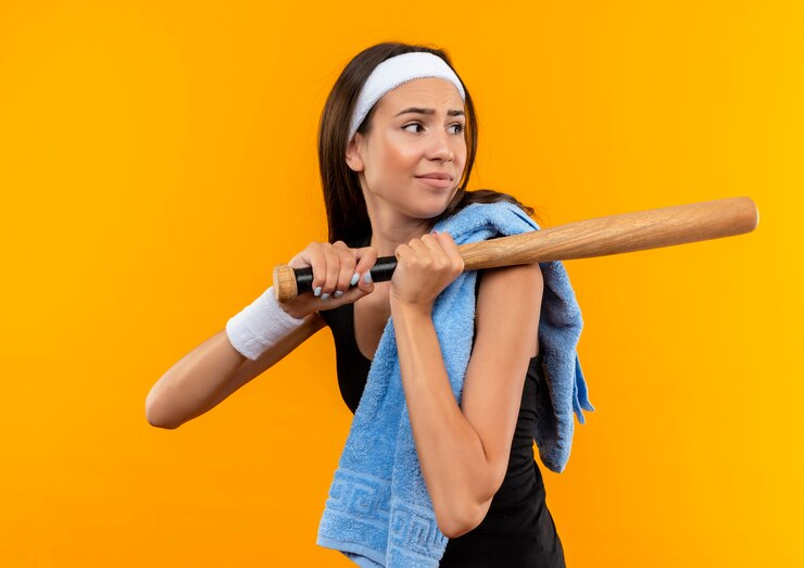 Foto grátis garota jovem e bonita e desagradável usando bandana e pulseira com uma toalha no ombro segurando o taco de beisebol e olhando para o lado isolado no espaço laranja