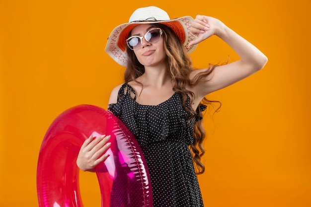 Garota jovem e bonita viajante descontente em um vestido de bolinhas com chapéu de verão usando óculos escuros segurando um anel inflável, olhando mostrando os polegares para baixo em pé sobre o espaço amarelo