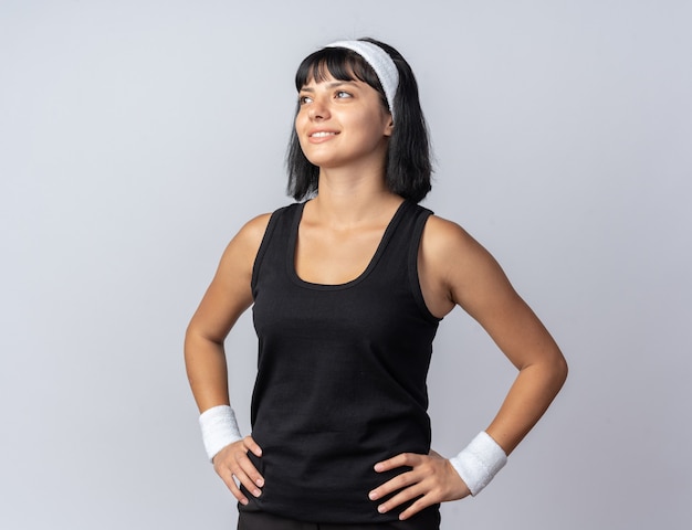 Garota jovem e feliz fitness usando bandana, olhando para cima e sorrindo alegremente em pé sobre um fundo branco