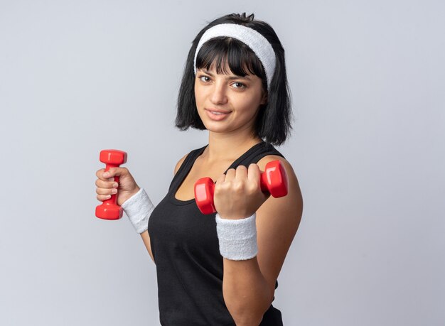 Garota jovem e feliz fitness usando uma bandana segurando halteres, fazendo exercícios, parecendo confiante em pé sobre um fundo branco