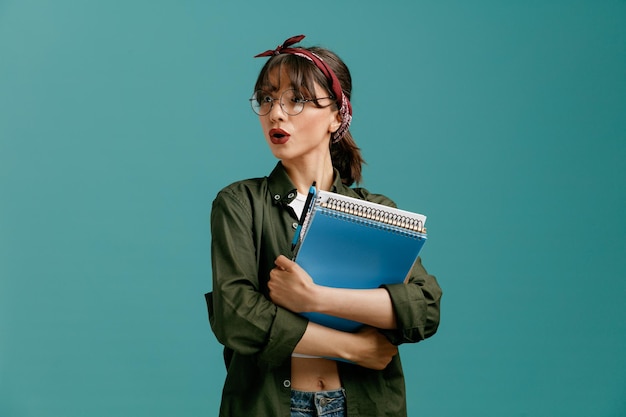 Garota jovem estudante surpresa usando óculos de bandana segurando grandes blocos de notas com caneta olhando para o lado enquanto abraça blocos de notas isolados em fundo azul