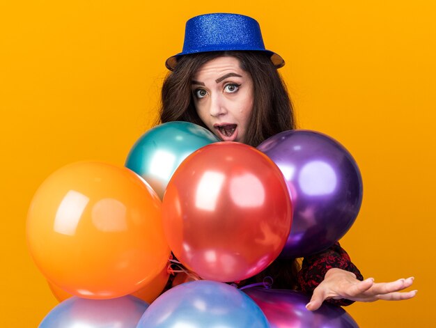 Garota jovem festeira com chapéu de festa em pé atrás de balões, olhando para a câmera, estendendo a mão isolada na parede laranja