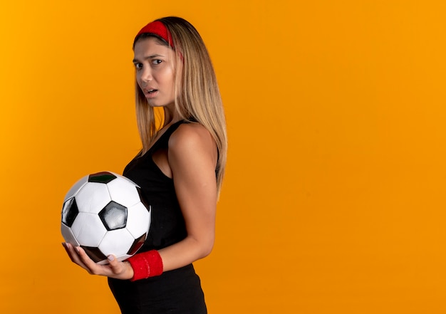 Foto grátis garota jovem fitness em roupa esportiva preta e bandana vermelha segurando uma bola de futebol descontente em pé sobre a parede laranja