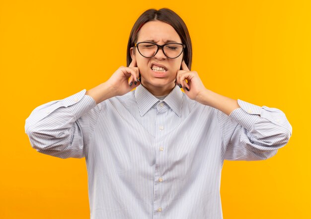 Garota jovem irritada do call center usando óculos, colocando os dedos nas orelhas com os olhos fechados, isolado em laranja