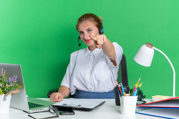 Garota jovem loira de call center satisfeita usando fone de ouvido, sentada na mesa com ferramentas de trabalho, olhando e apontando para a câmera isolada na parede verde