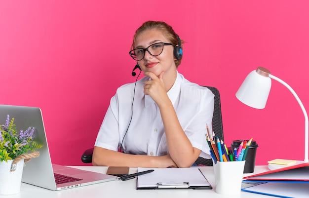 Garota jovem loira satisfeita com o call center usando fone de ouvido e óculos, sentada na mesa com ferramentas de trabalho, segurando o queixo, olhando para a câmera isolada na parede rosa