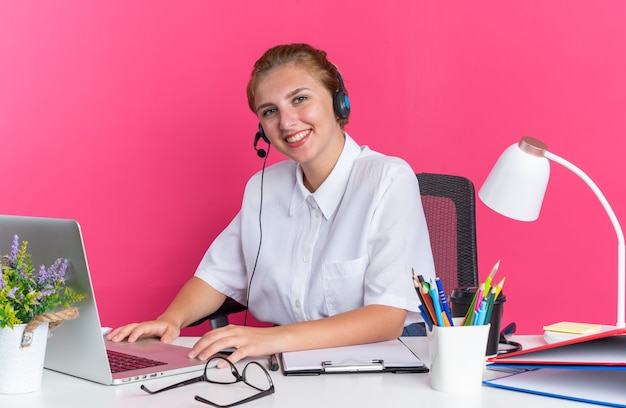 Garota jovem loira sorridente de call center usando fone de ouvido, sentada na mesa com ferramentas de trabalho, usando laptop