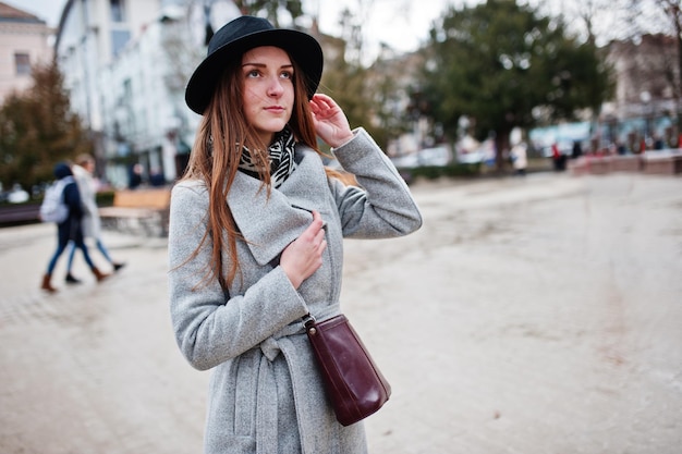 Garota jovem modelo turista em um casaco cinza e chapéu preto com bolsa de couro nos ombros posou na rua da cidade