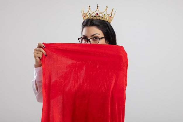 Foto grátis garota jovem super-heroína caucasiana confiante usando óculos e uma coroa segurando sua capa de herói, cobrindo o rosto por trás dela, isolada na parede branca com espaço de cópia