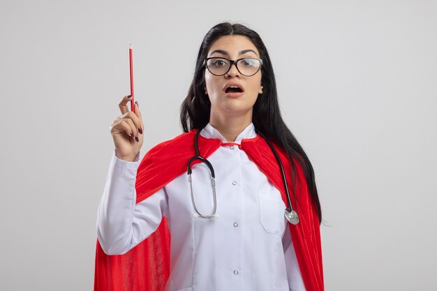 Garota jovem super-heroína caucasiana impressionada usando óculos e estetoscópio segurando um lápis vermelho, olhando para a câmera isolada no fundo branco com espaço de cópia