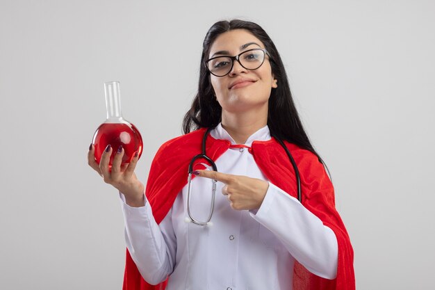 Garota jovem super-heroína caucasiana satisfeita usando óculos e estetoscópio segurando e apontando para o frasco químico com um líquido vermelho, olhando para a câmera isolada no fundo branco