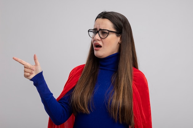 Garota jovem super-heroína confusa usando óculos, olhando e apontando para o lado isolado no fundo branco