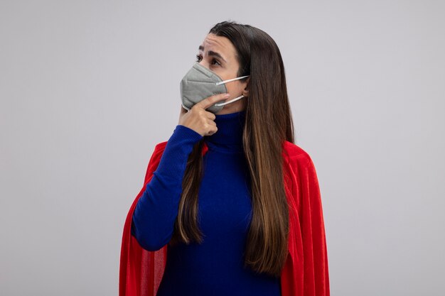 Garota jovem super-heroína impressionada olhando para o lado usando máscara médica e colocando a mão sob o queixo isolado no branco