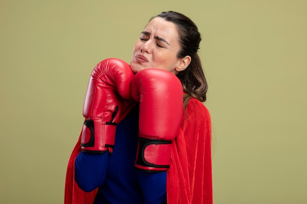 Garota jovem super-heroína insatisfeita com os olhos fechados e luvas de boxe e colocar as mãos sob o queixo isolado em fundo verde oliva