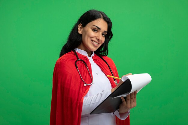 Garota jovem super-heroína sorridente, olhando para a frente, usando um manto médico com um estetoscópio segurando e escrevendo algo na área de transferência isolada em verde