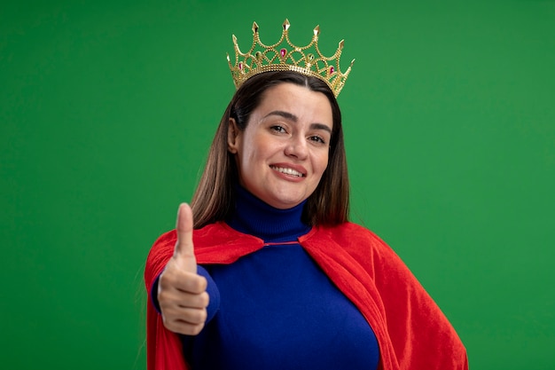 Garota jovem super-heroína sorridente usando uma coroa aparecendo o polegar isolado no fundo verde