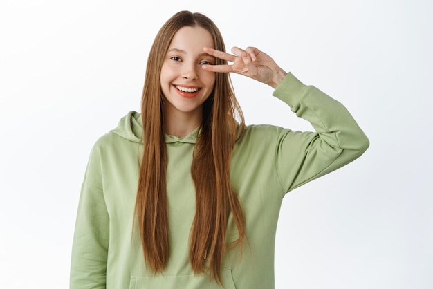 Garota kawaii positiva com cabelo comprido mostra gesto de paz vsign sorrindo otimista em pé sobre fundo branco Copiar espaço