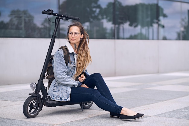 Foto grátis garota legal pensativa de óculos e com tatuagens no pescoço e nas mãos está relaxando em sua própria scooter.