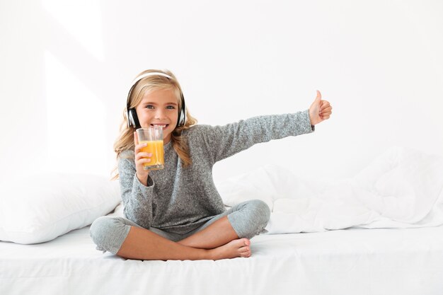 Garota linda criança em fones de ouvido, segurando o copo de suco de laranja, mostrando o polegar para cima gesto, enquanto está sentado na cama