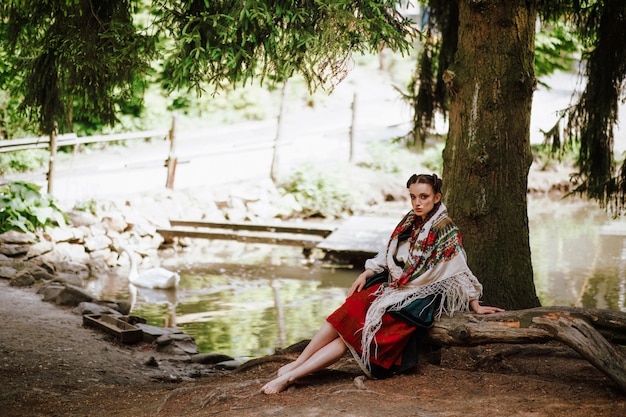 Foto grátis garota linda em um vestido bordado ucraniano, sentado em um banco perto do lago