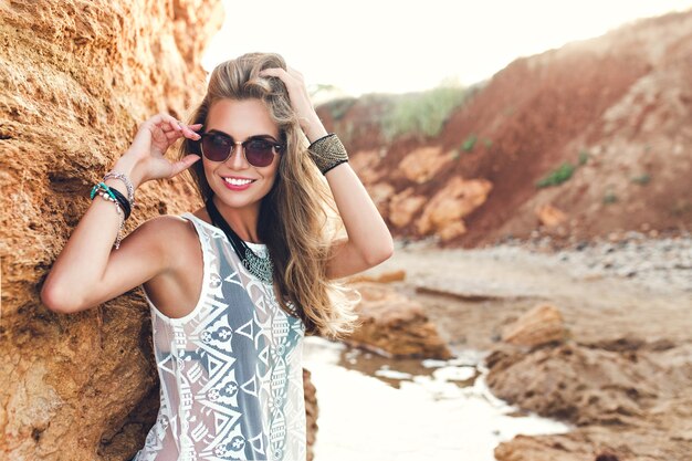 Garota loira atraente com cabelo comprido está posando para a câmera em fundo de pedras. Ela está tocando o cabelo e sorrindo.