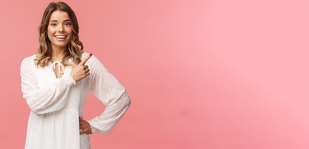 Foto grátis garota loira caucasiana otimista e bonita de vestido branco, apontando o canto superior direito e sorrindo para a câmera com um rosto curioso interessado discutir descontos especiais de primavera promocionais fundo rosa