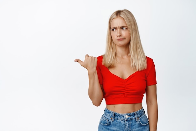 Garota loira cética apontando e olhando para a esquerda com cara de mau humor expressa dúvida ou antipatia em pé no top recortado e jeans contra fundo branco