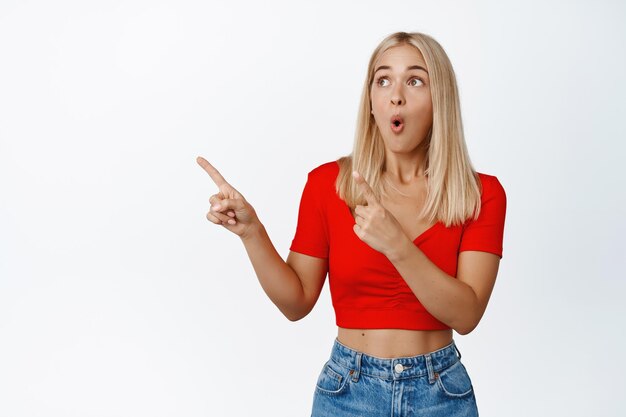 Garota loira espantada verificando o grande anúncio na loja apontando dedos e olhando para a esquerda no anúncio sobre fundo branco
