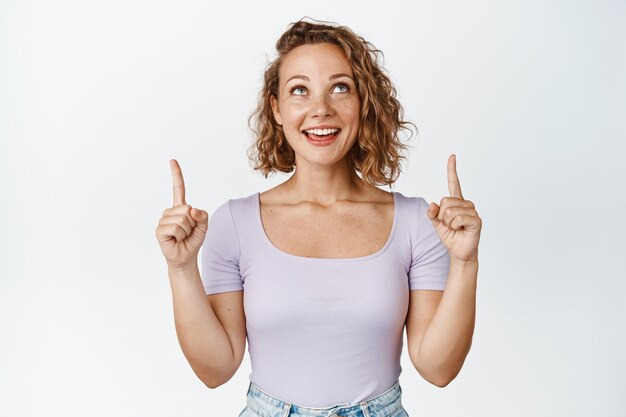 Garota loira feliz apontando os dedos para cima e olhando para a oferta promocional, sorrindo, alegre, mostrando um bom negócio, anúncio de venda em branco