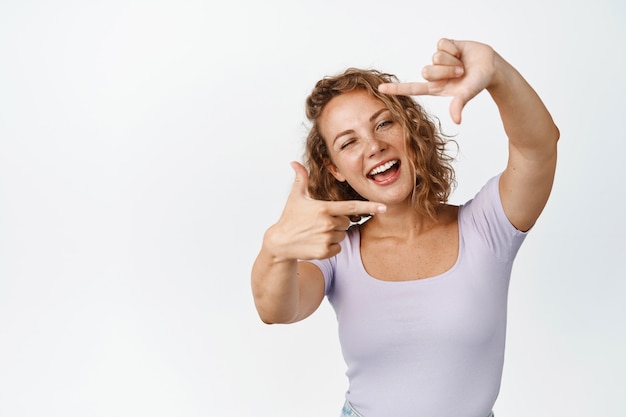 Garota loira feliz olhando através de quadros de mão com um sorriso alegre, retratando o momento, tirando um gesto de imagem, de pé no branco.