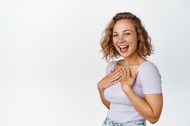 Garota loira feliz se divertindo, rindo e sorrindo com alegria, segurando as mãos no peito e assistindo algo engraçado em branco.