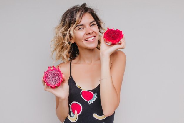 Garota loira na moda posando com frutas exóticas. Tiro interno de entusiasmada senhora caucasiana em vestido segurando pitaya.