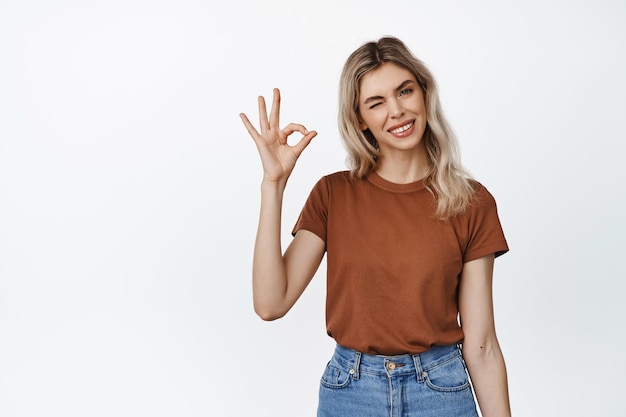 Garota loira positiva piscando mostrando sinal de OK OK para aprovar produto recomendado ou loja em pé em camiseta e jeans contra fundo branco