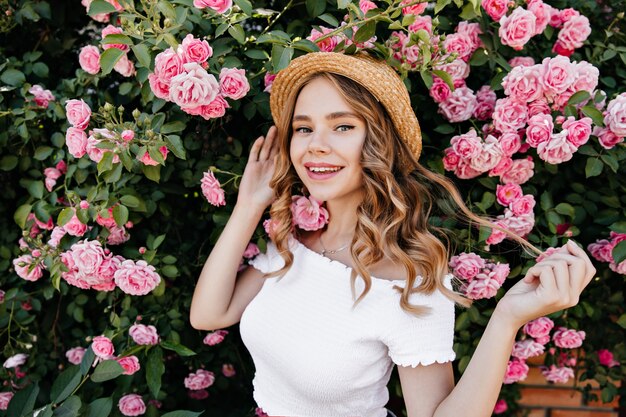 Garota loira romântica posando com um sorriso na frente de lindas flores. Retrato ao ar livre de alegre mulher cacheada brincando com seu cabelo no jardim.