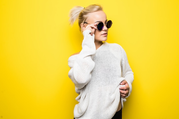 Garota loira séria da moda com um suéter branco moderno e óculos de sol azuis brilhantes posando em amarelo