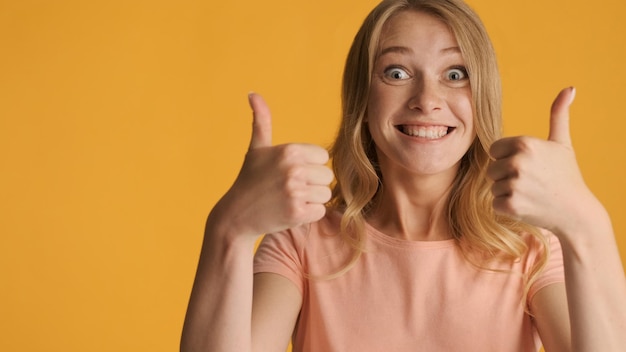 Foto grátis garota loira sorridente animada mantendo os polegares para cima perto com espaço de cópia para texto ou conteúdo promocional isolado em fundo amarelo como expressão