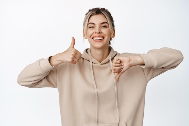 Garota loira sorridente com dentes brancos mostrando os polegares para cima e para baixo como ou não gosta de gesto em pé contra um fundo branco Copiar espaço