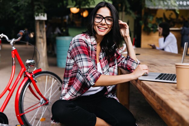 Garota maravilhosa de bom humor, sentado na cidade com o laptop e sorrindo. Retrato ao ar livre de uma senhora morena atraente em copos, posando ao lado da bicicleta.