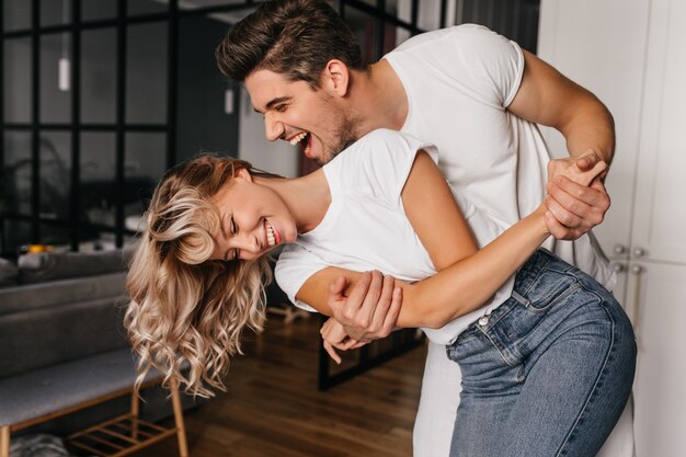 Garota maravilhosa em t-shirt branca dançando com o namorado. Retrato interno de senhora cativante brincando em casa com o marido.