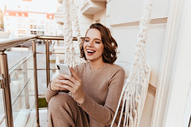 Garota maravilhosa lendo mensagem de telefone enquanto está sentado no terraço. Foto de animada senhora bonita posando com smartphone na varanda.