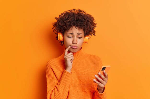 Garota milenar chateada com expressão facial frustrada lê mensagem de texto ouve música da lista de reprodução vestida com um suéter casual isolado sobre uma parede laranja viva