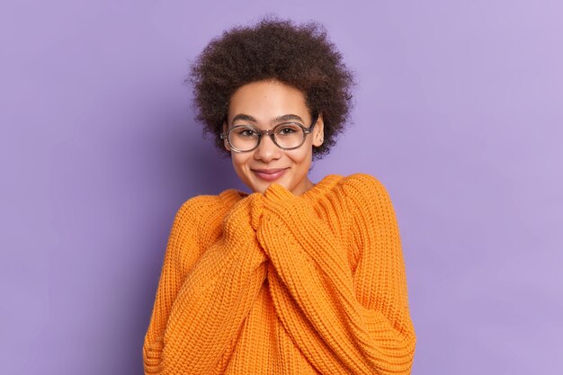 Garota milenar de cabelos muito encaracolados mantém as mãos perto de sorrisos no queixo e ouve algo bom usa óculos óticos, suéter laranja de malha