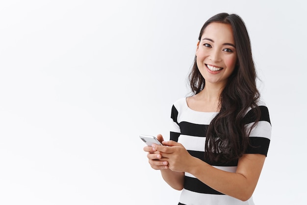 Garota morena do leste, tenra e sorridente, de pé, virada pela metade, sobre fundo branco, com roupa casual, escrevendo mensagem via smartphone, convidando amigos, festa de halloween, olhando a câmera alegremente