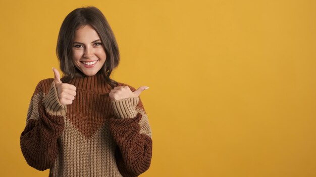 Garota morena sorridente atraente em suéter de malha aconchegante mostrando alegremente os polegares para cima gestos sobre fundo colorido