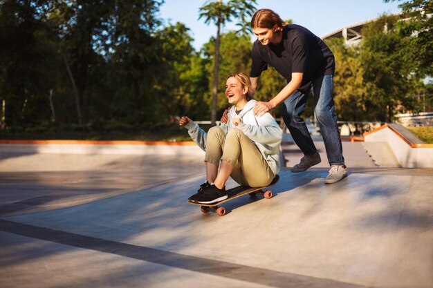 Garota muito alegre sentada no skate e andando com cara jovem perto enquanto alegremente passando tempo juntos no skatepark