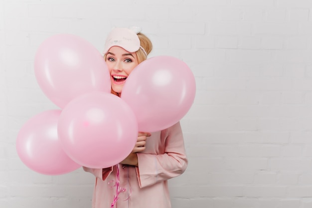 Garota muito emocional de pijama segurando balões de festa pela manhã. Mulher loira excitada comemorando algo, rindo na parede branca.